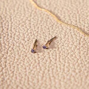 small dark purple stud earrings photographed on a textured cream surface beside a fine gold chain